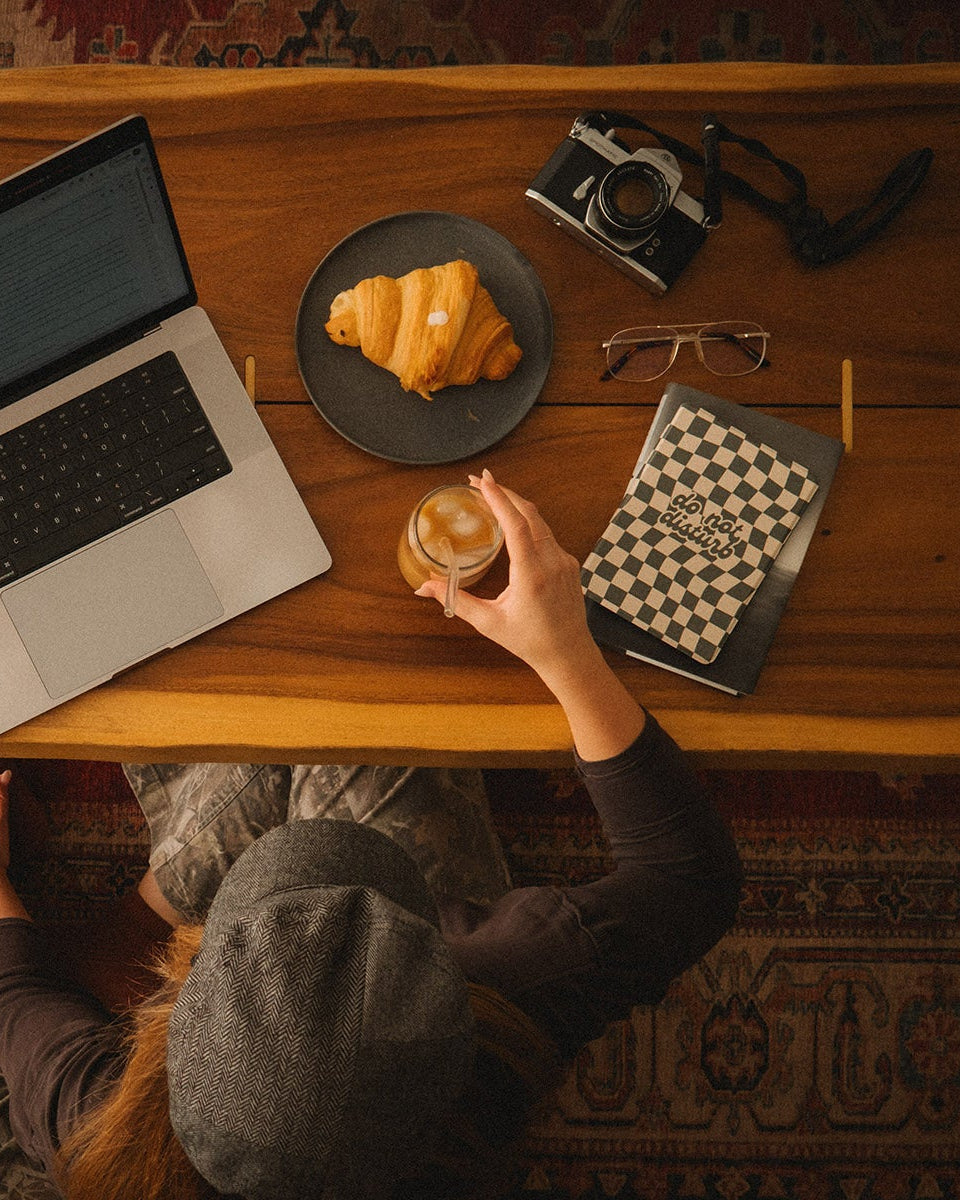 Person sitting at a wooden table with a laptop, croissant, and camera.