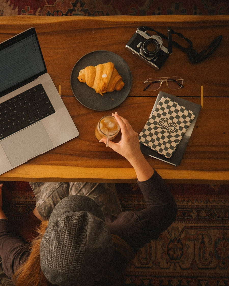 Person sitting at a wooden table with a laptop, croissant, and camera.